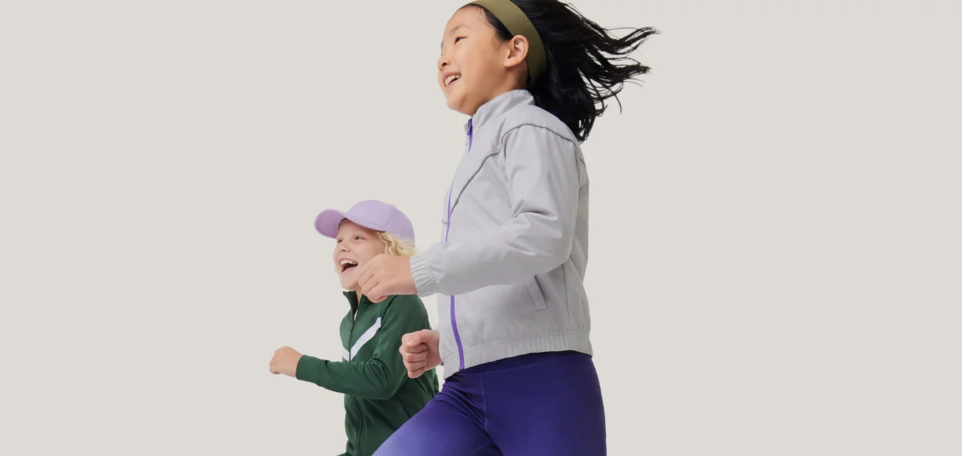 Due bambini corrono fianco a fianco, indossando abbigliamento sportivo e scarpe da ginnastica, una ragazza con giacca e leggings e l'altra con cappellino e tuta.