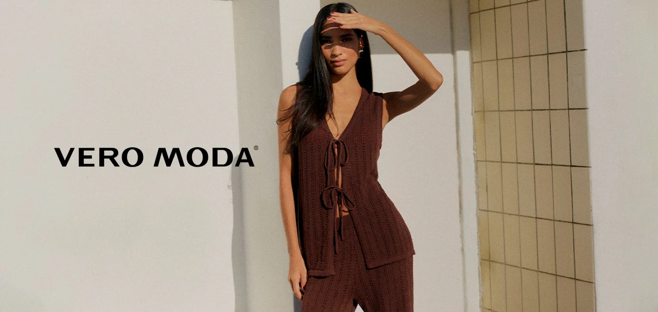 Donna con top marrone lavorato a maglia senza maniche e pantaloni in piedi scalza su un patio soleggiato, protegge gli occhi con la mano contro il cielo azzurro chiaro.