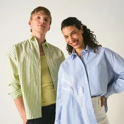 Due persone stanno sorridenti contro uno sfondo semplice, indossando camicie larghe a righe e pantaloni casual, posando con sicurezza.