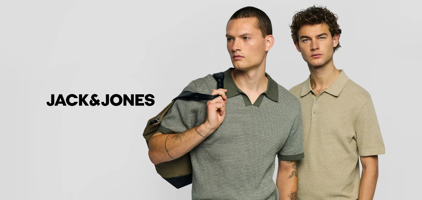 Due uomini stanno fianco a fianco indossando polo in maglia a maniche corte, uno grigio con colletto verde e borsa a tracolla, l’altro beige con le mani in tasca.