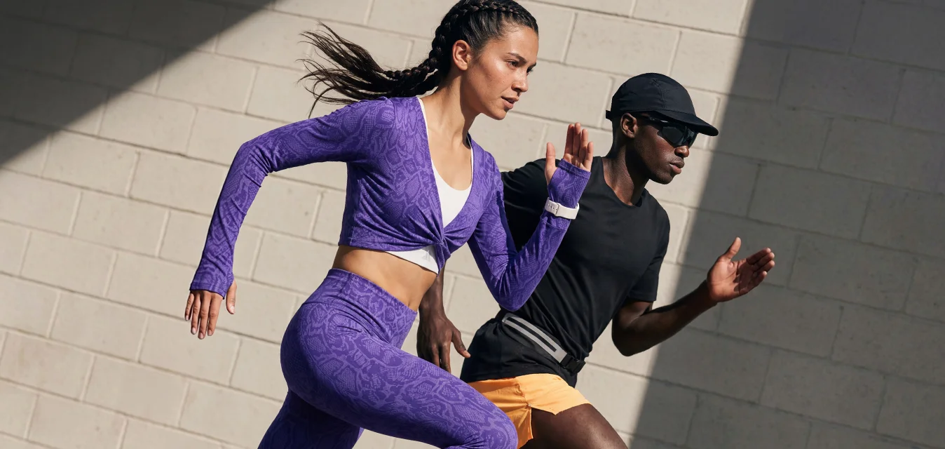 Due corridori, una donna con abbigliamento sportivo viola e un uomo con abbigliamento sportivo nero e giallo, corrono a tutta velocità all'aperto davanti a un muro di mattoni grigi.