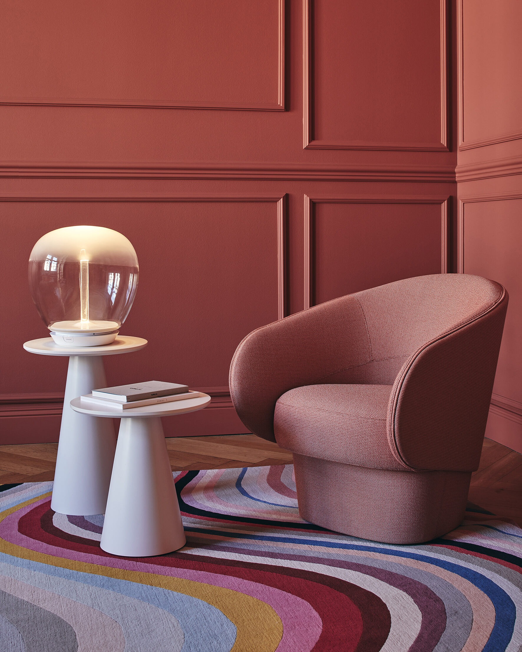 Textured mauve accent armchair beside white pedestal tables with glass globe lamp, books and colourful wavy rug on parquet floor