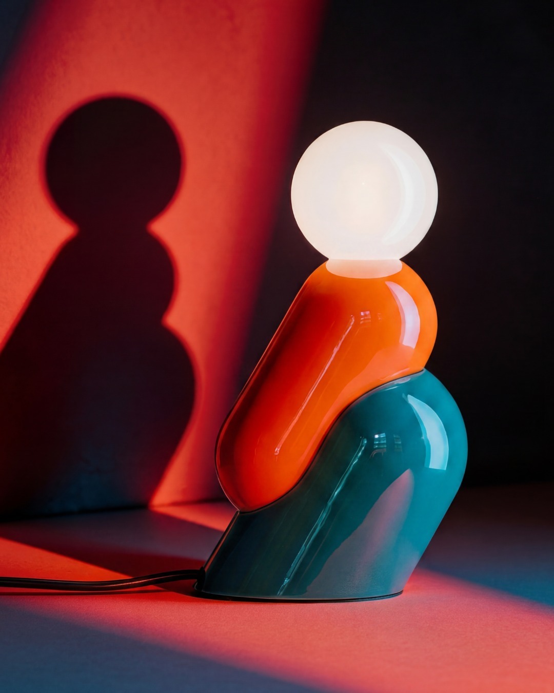 Colourful modern table lamp with round white bulb and glossy orange-and-teal sculptural base casting a dramatic red shadow