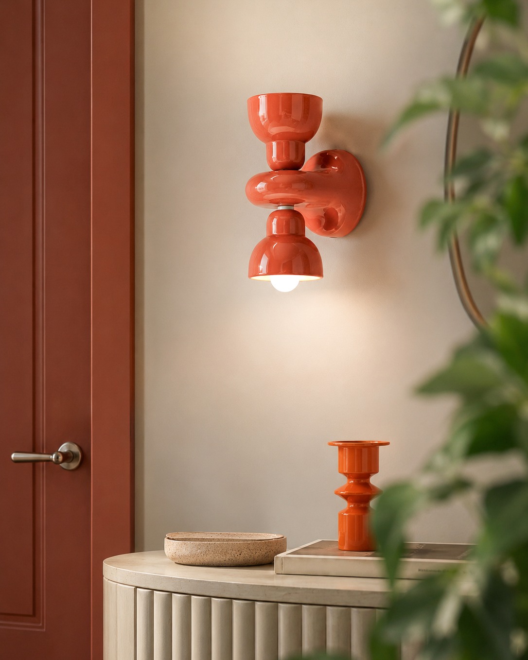 Coral ceramic wall sconce lit above a cream console table with matching coral candle holder, red door and greenery