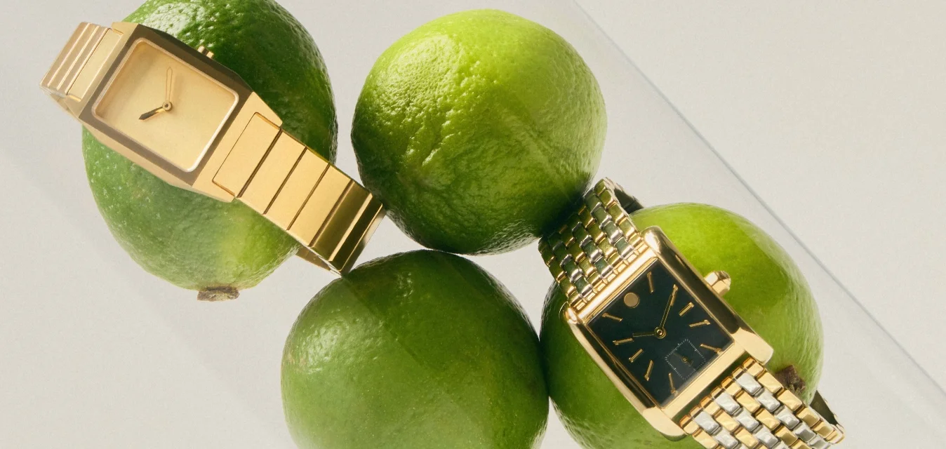 Sei lime verdi all'interno di un cilindro di vetro trasparente con due orologi da polso dorati posati sopra le lime.