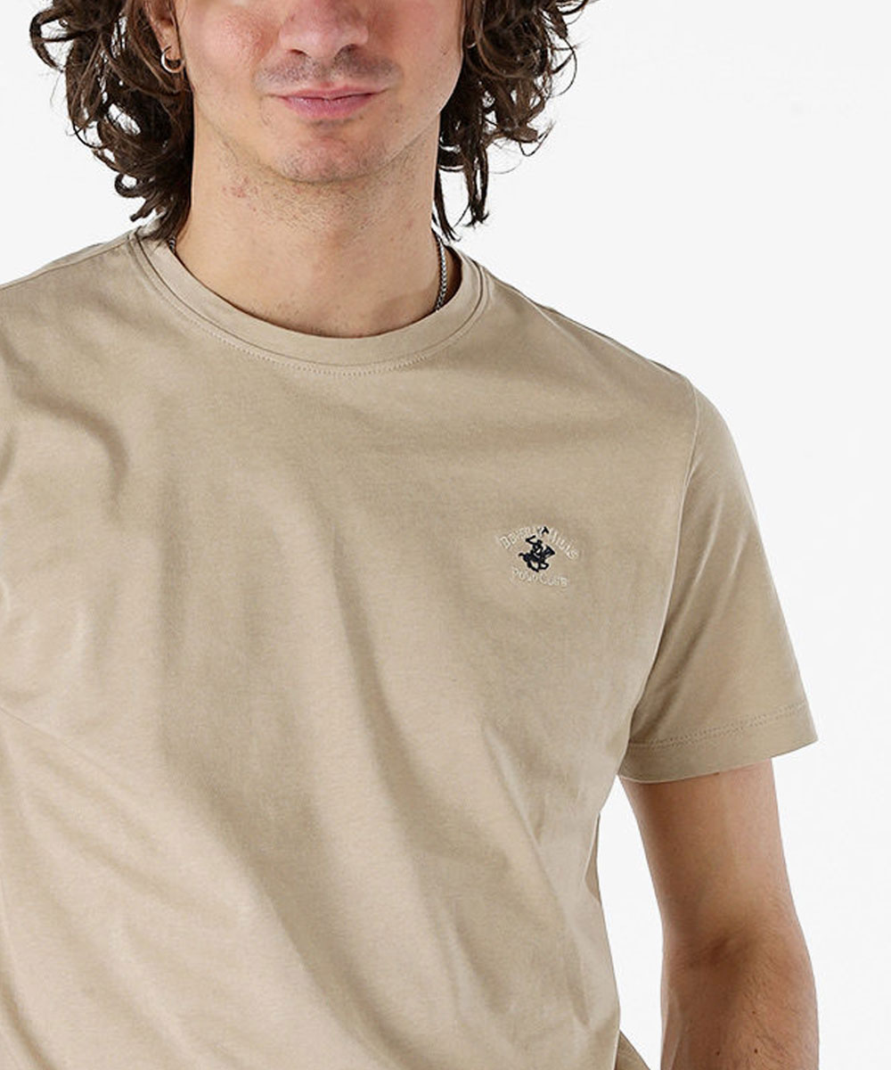 Young man wearing beige short-sleeve t-shirt with small embroidered logo on left chest, curly hair and silver necklace