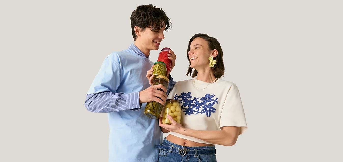 Giovane uomo e donna sorridenti, che tengono barattoli di verdure sottaceto, in piedi contro uno sfondo chiaro e semplice.