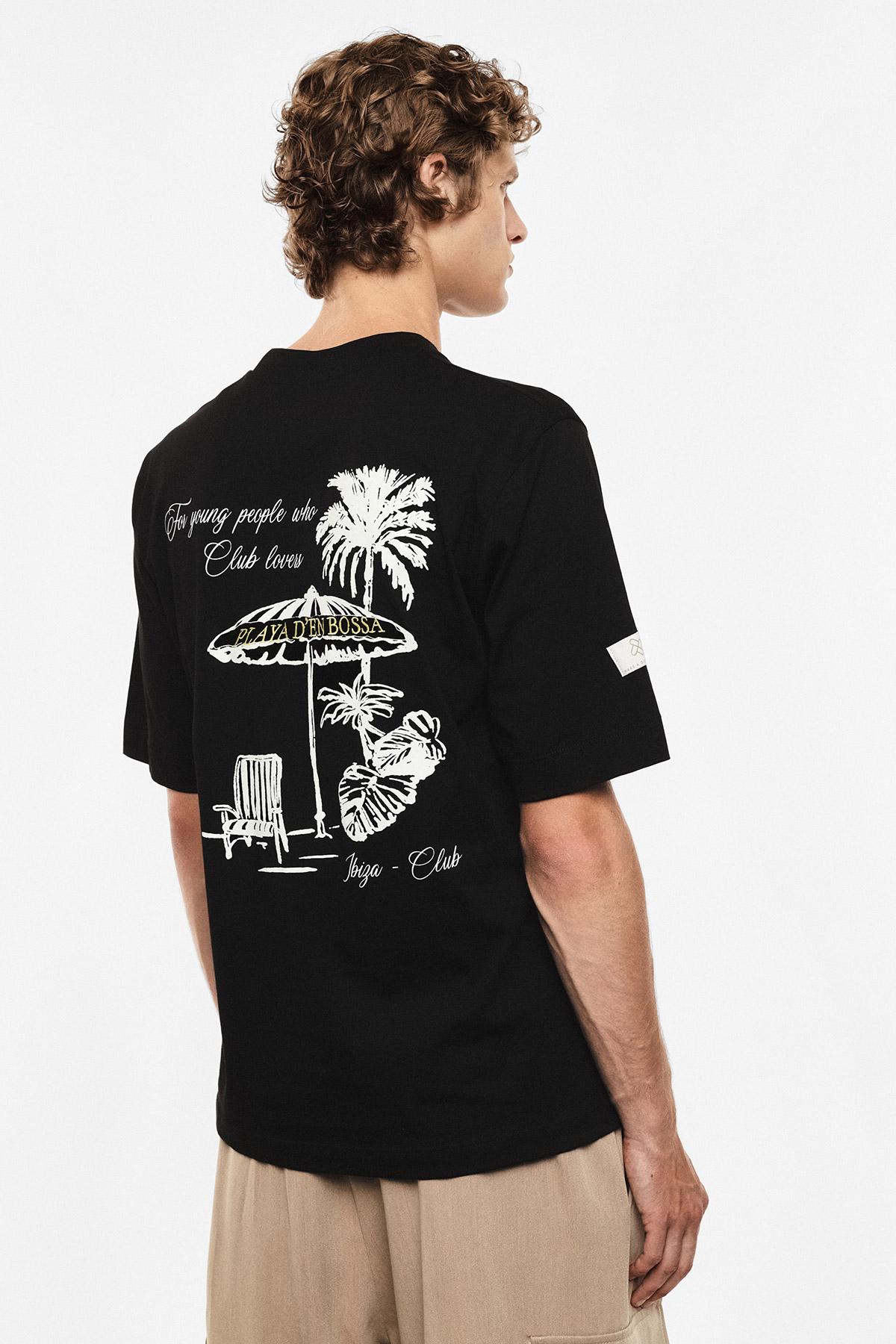Back view of a person wearing a black T-shirt with white palm tree and beach umbrella graphic and beige trousers