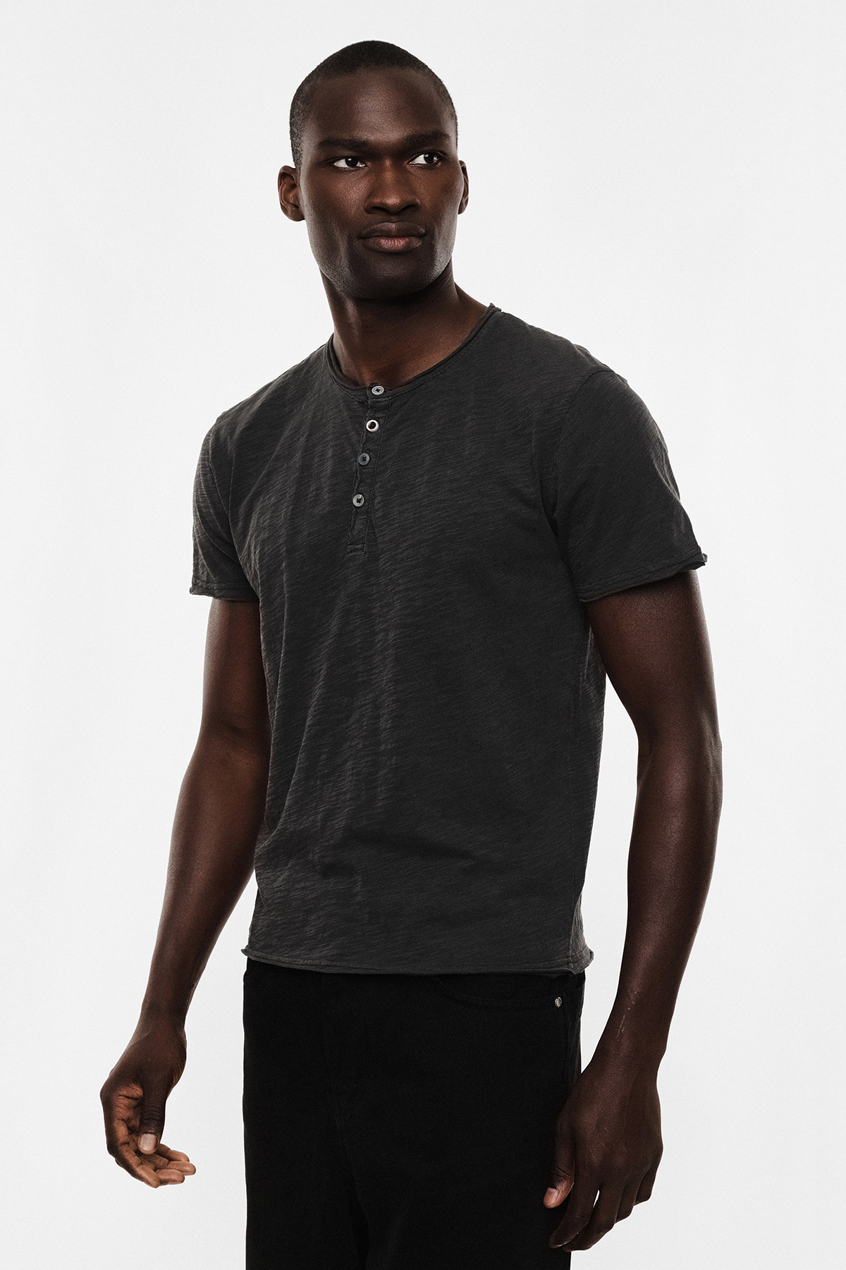 Male model wearing a charcoal henley T-shirt and black trousers, standing against a white background and looking to his left