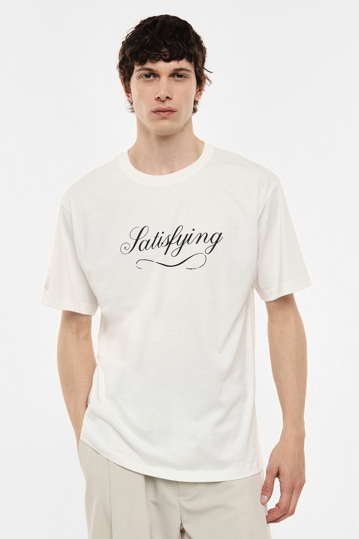 Man wearing white T-shirt printed with 'Satisfying' script and beige trousers, standing against a plain white background