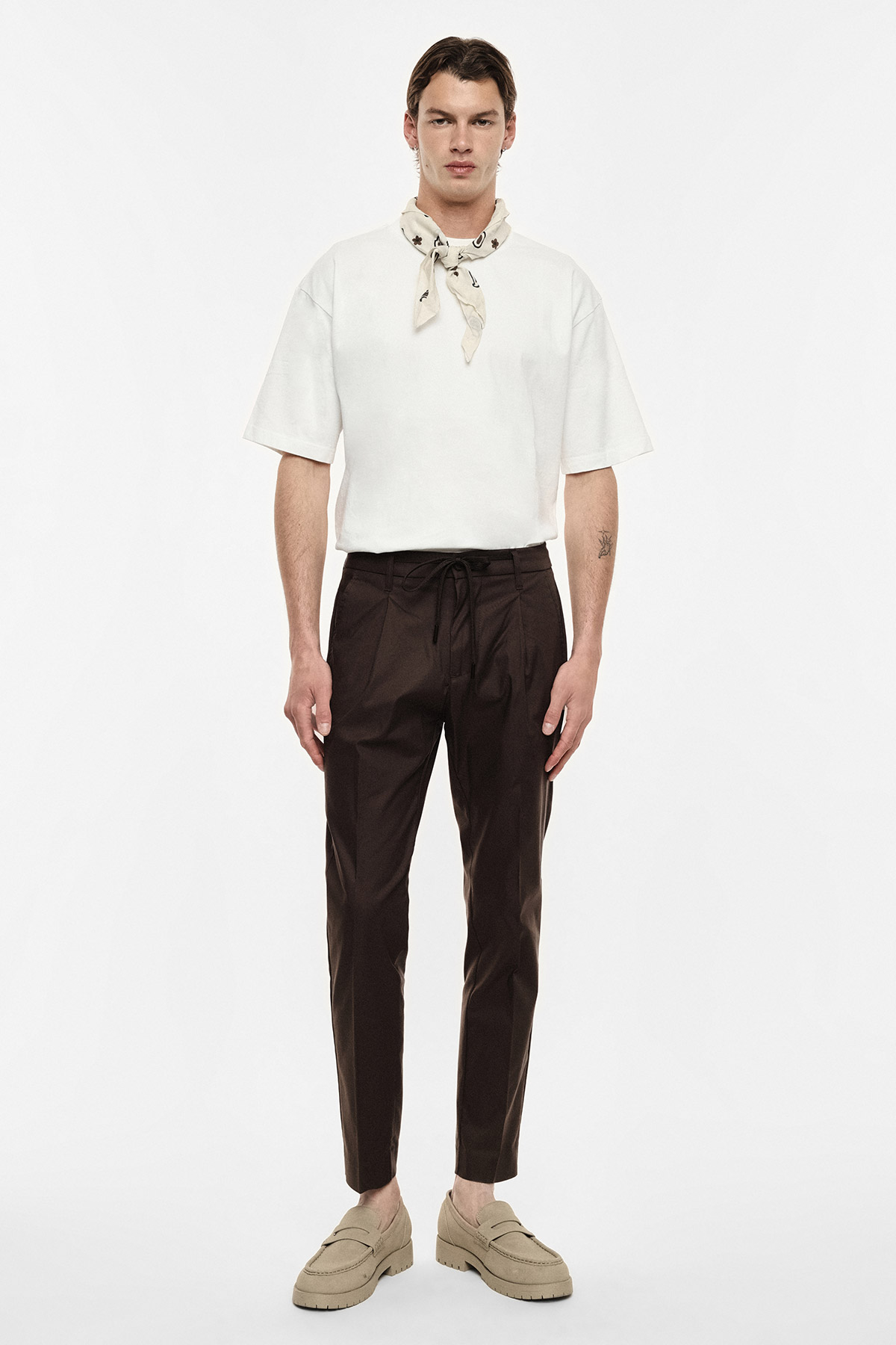 Man wearing white boxy T-shirt, beige neckerchief, brown tapered trousers and beige loafers, standing full‑length