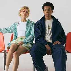 Due persone sono sedute su sedie di plastica arancioni. Una indossa una giacca a righe verdi e pantaloni corti; l'altra, una tuta blu navy. Sono visibili scarpe da ginnastica e calze.