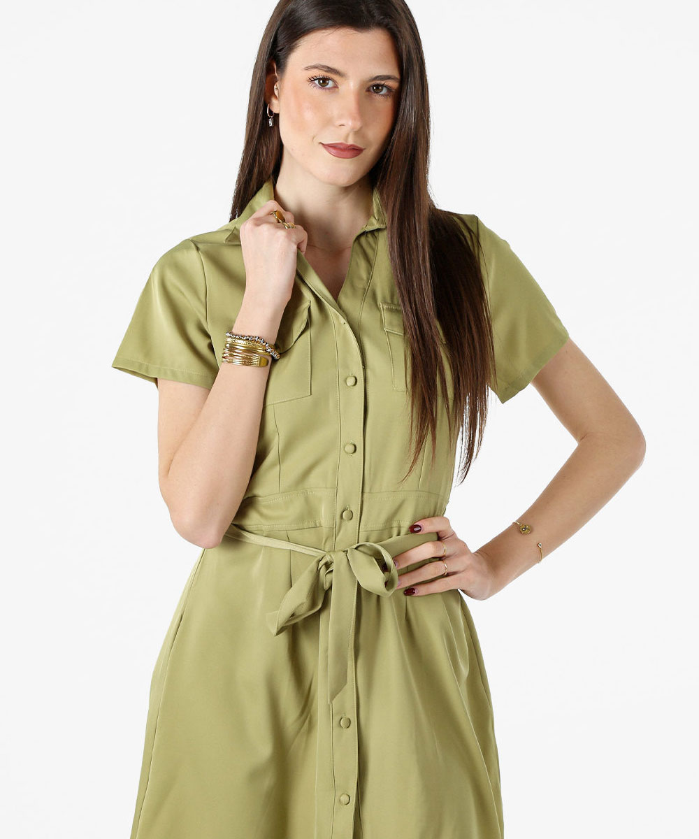 Woman modelling an olive-green short-sleeve button-front shirt dress with tie waist, long straight hair and bracelets on a white background