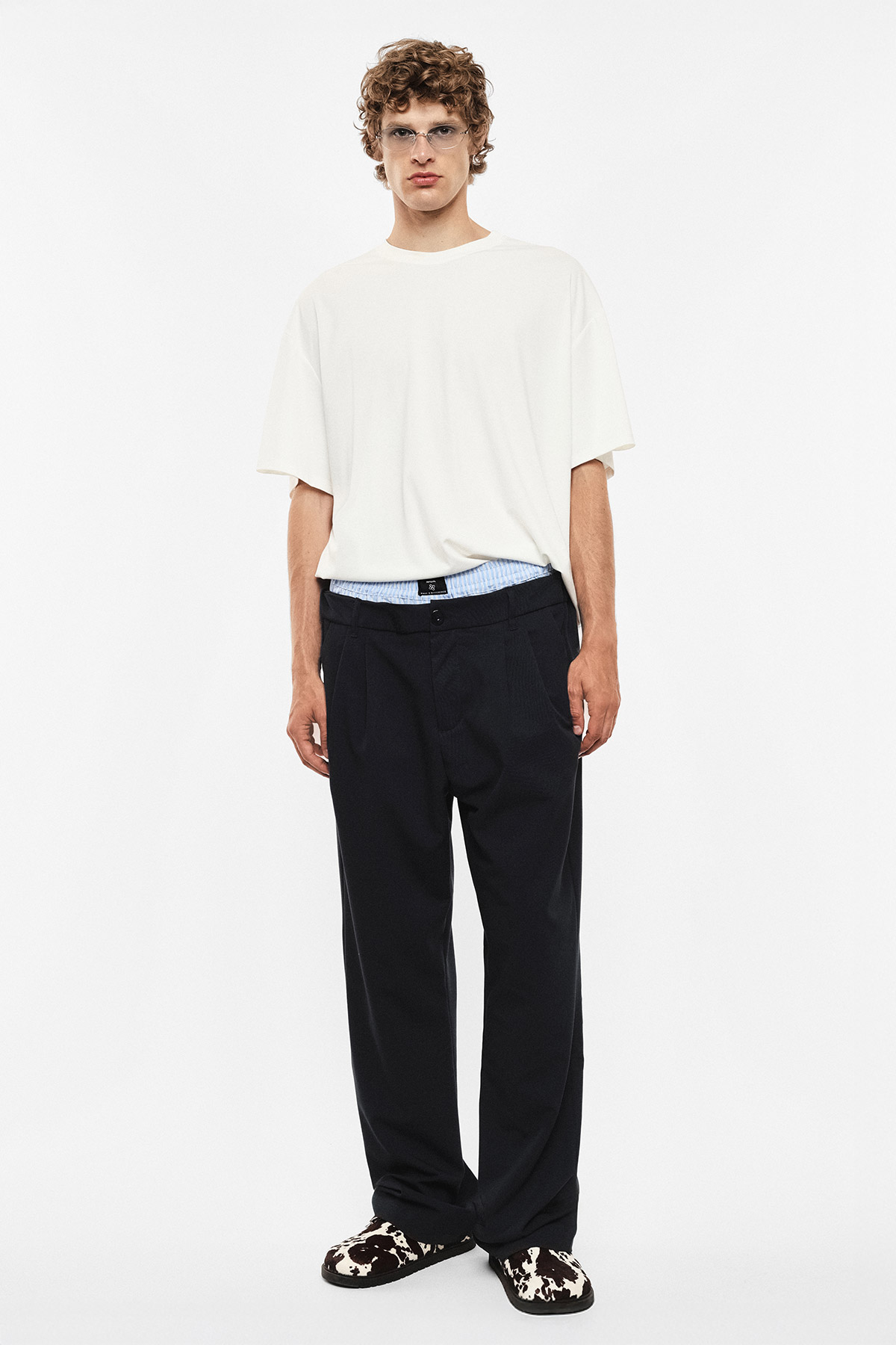 Male model wearing oversized white T-shirt, navy trousers and patterned slippers, standing front-on against white background