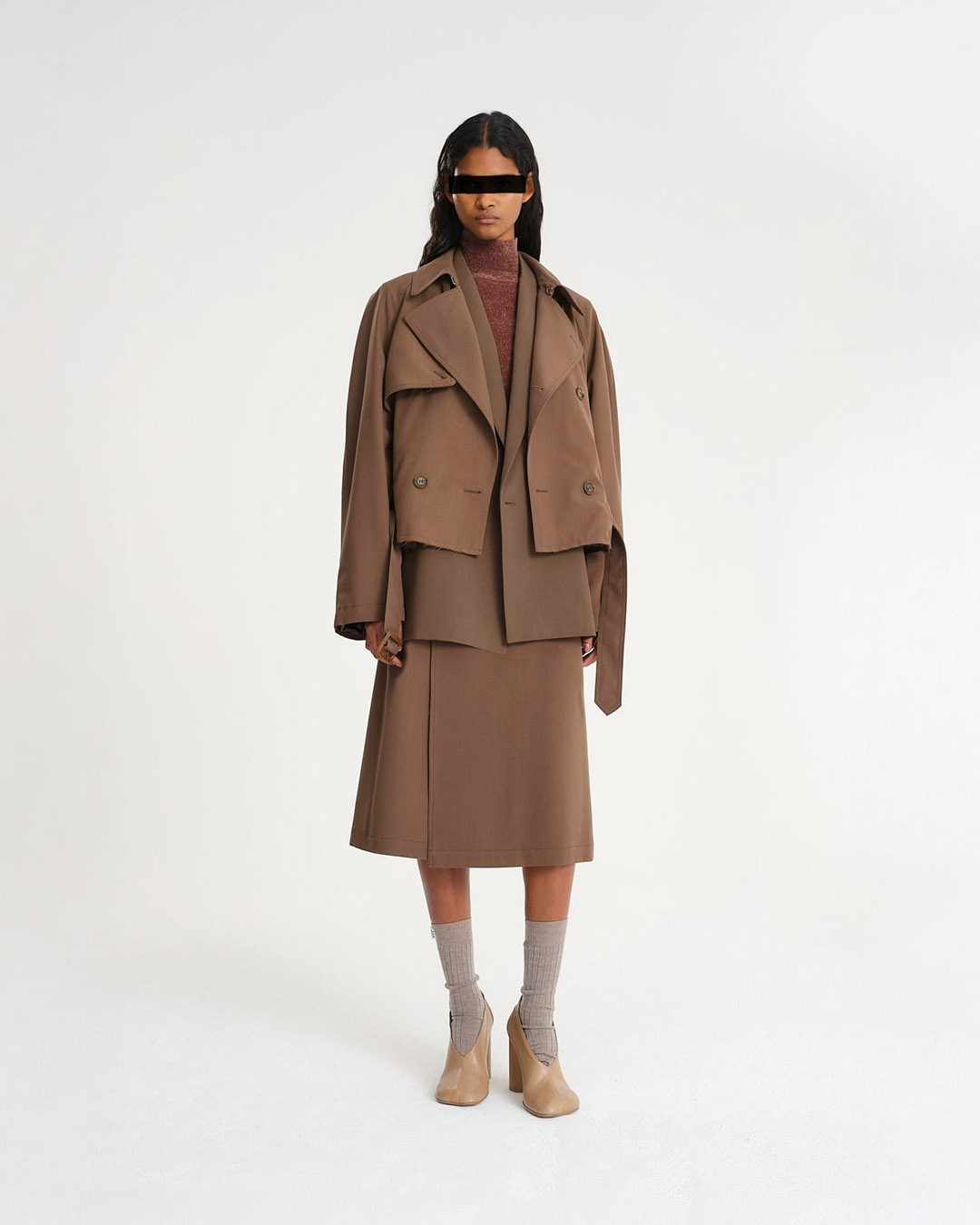 Model wearing brown layered tailored outfit: cropped trench jacket, blazer, midi skirt and ankle boots with socks on white background