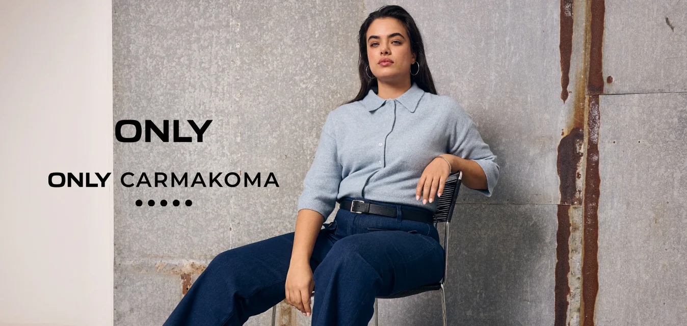 Donna con i capelli lunghi seduta su una sedia, indossa un maglione azzurro, pantaloni blu scuro e mocassini marroni, davanti a una parete industriale grigia.