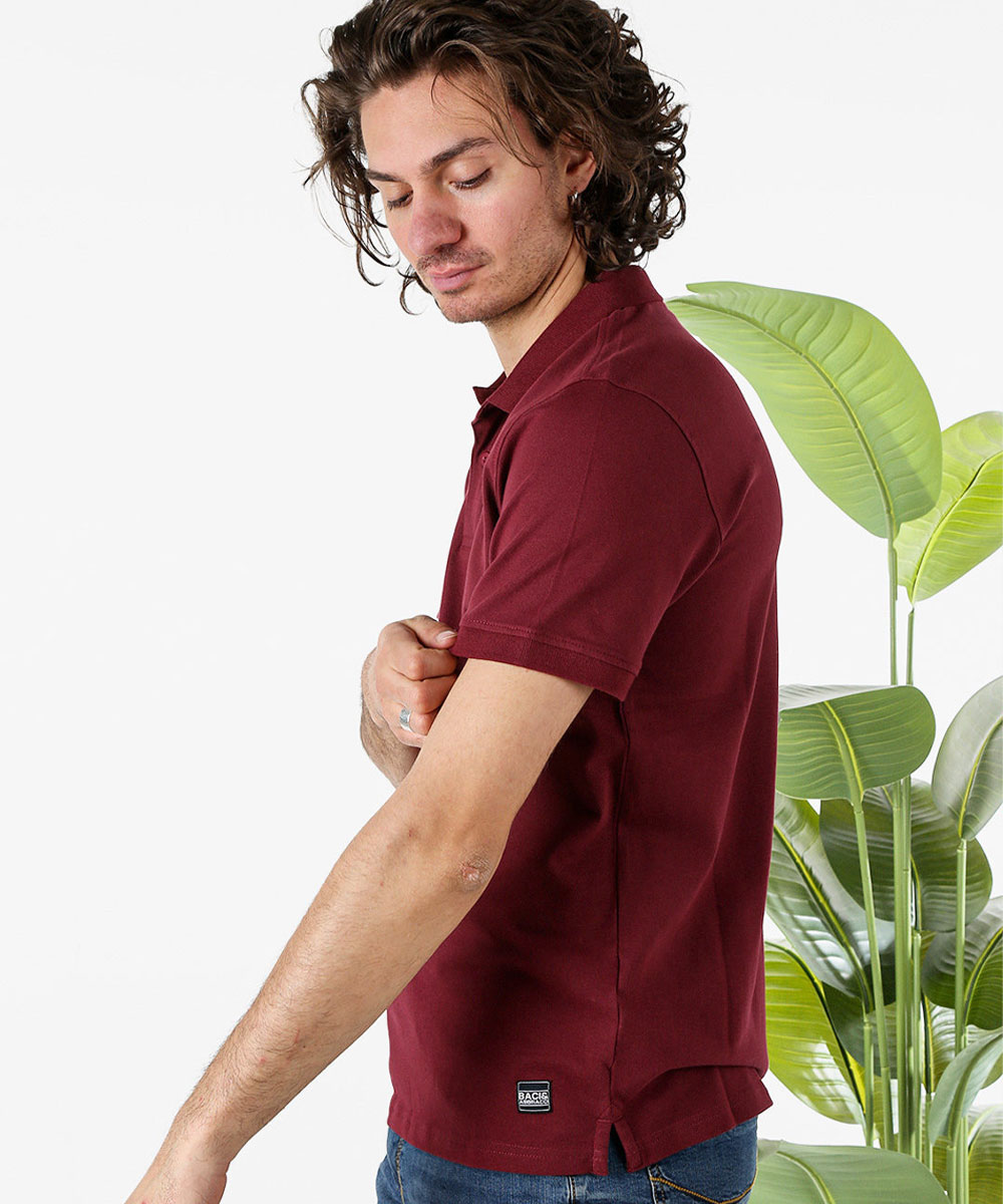 Person adjusting sleeve of burgundy polo shirt in side view, wearing jeans with green houseplant background, casual apparel