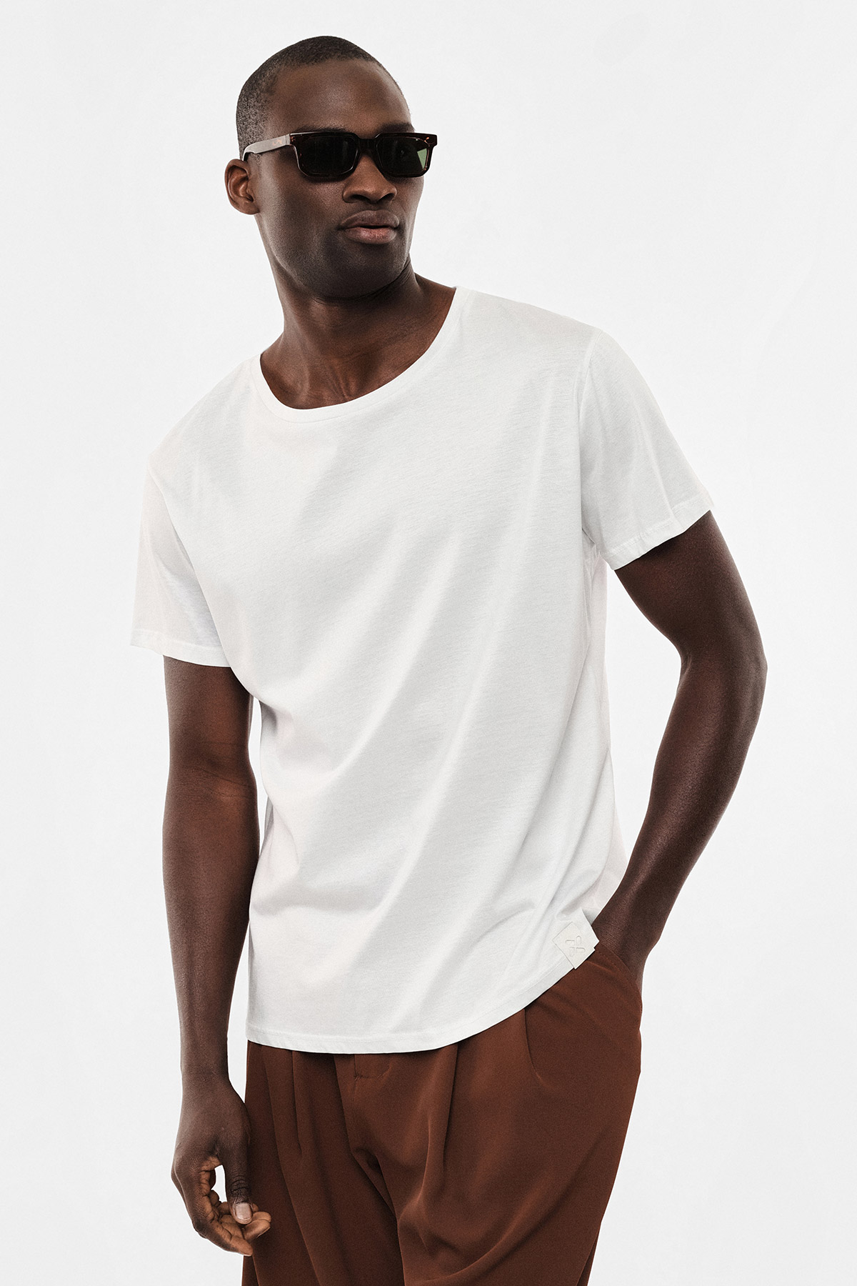 Male model wearing plain white T-shirt, brown trousers and sunglasses, casual pose against white background