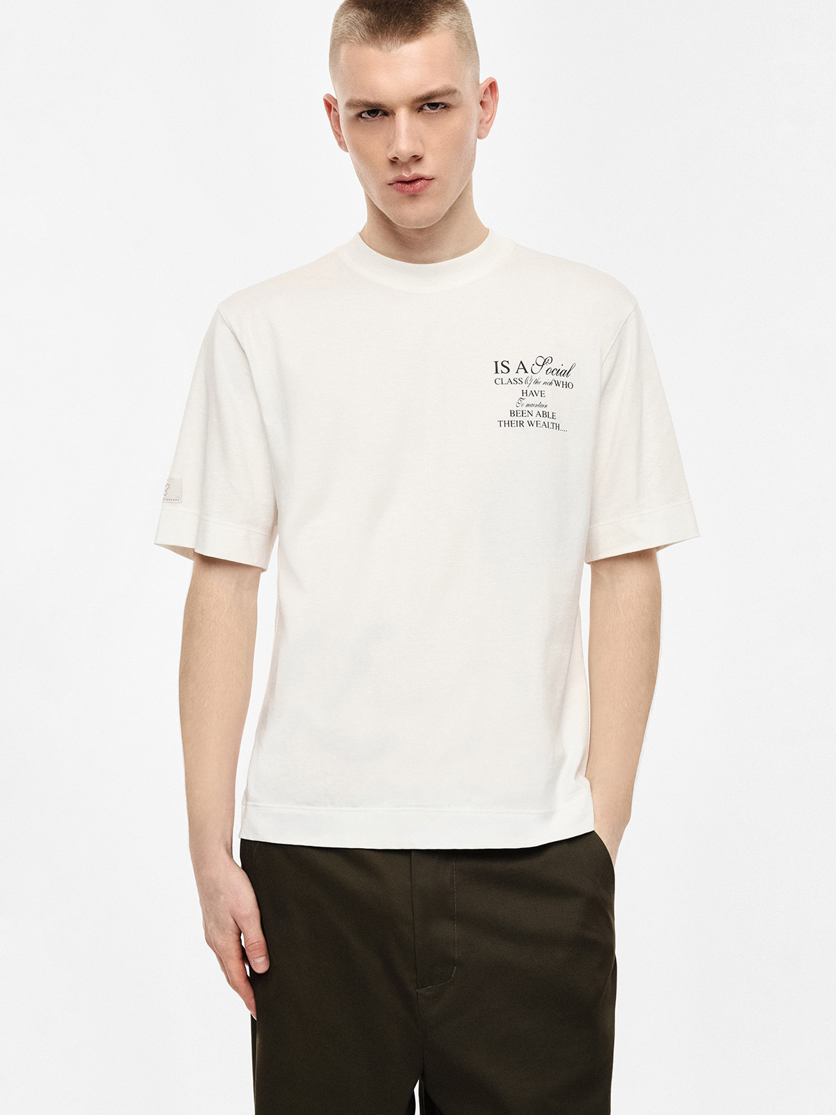 Male model wearing white short-sleeve T-shirt with small black chest text print and dark trousers on plain studio background