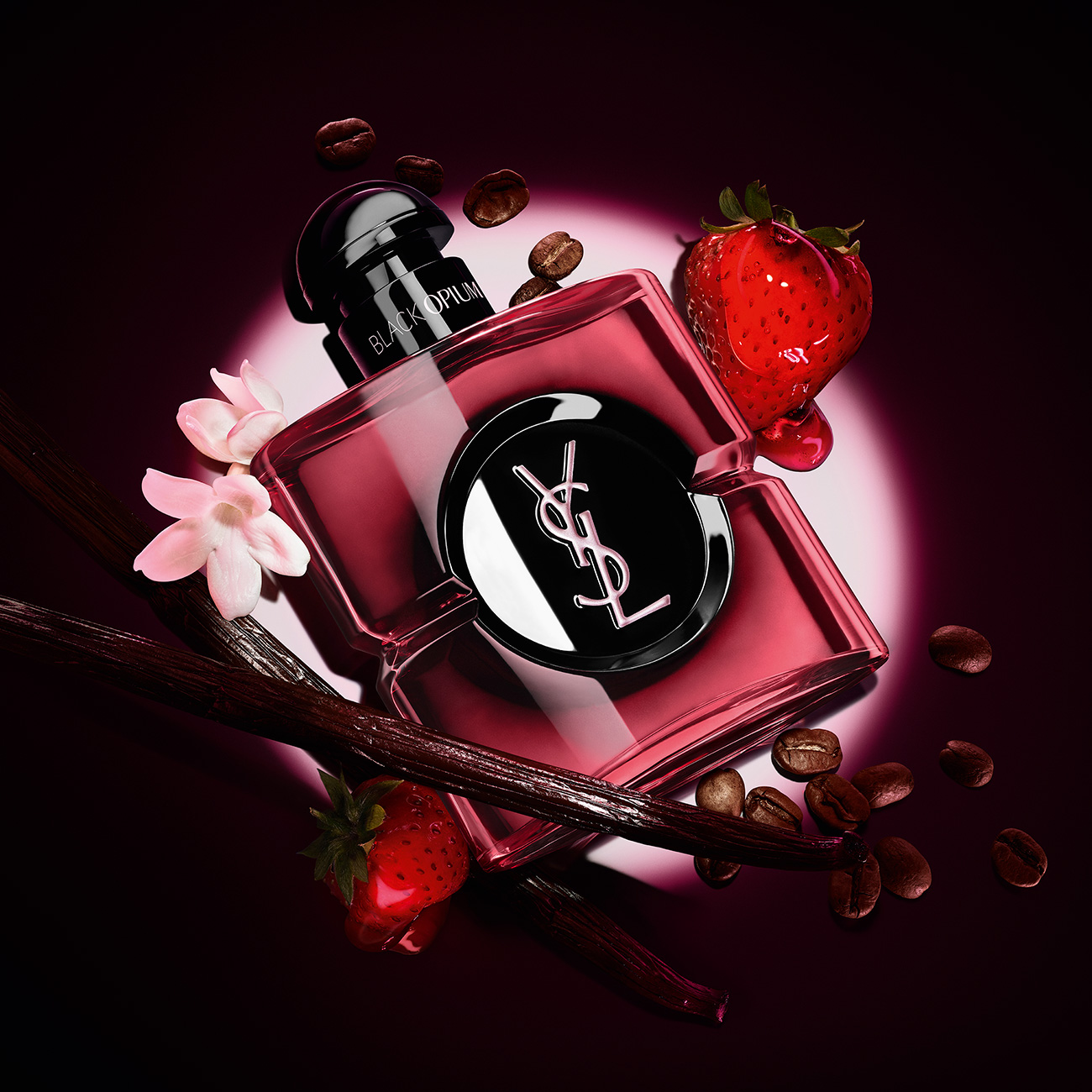 Un flacone rosso ciliegia traslucido del profumo YSL Black Opium circondato da chicchi di caff&egrave;, baccelli di vaniglia, fragole rosse e fiori bianchi sotto un riflettore.