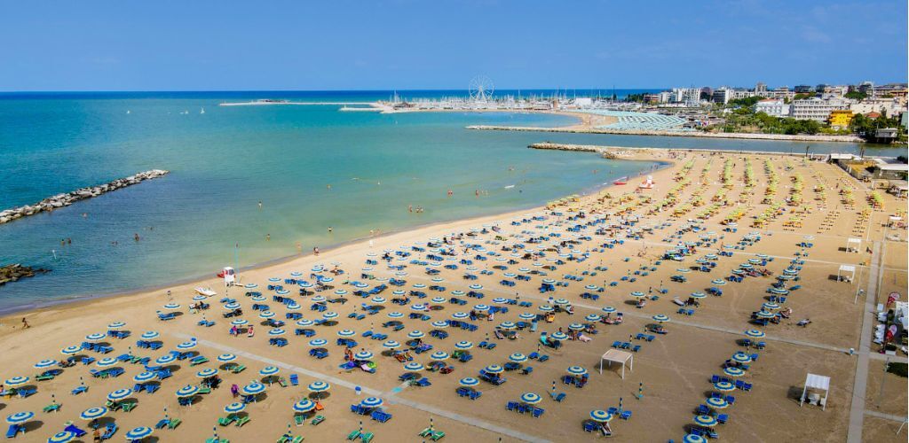 Aerial view of sandy Mediterranean beach with rows of blue-and-white umbrellas, turquoise sea and seaside town