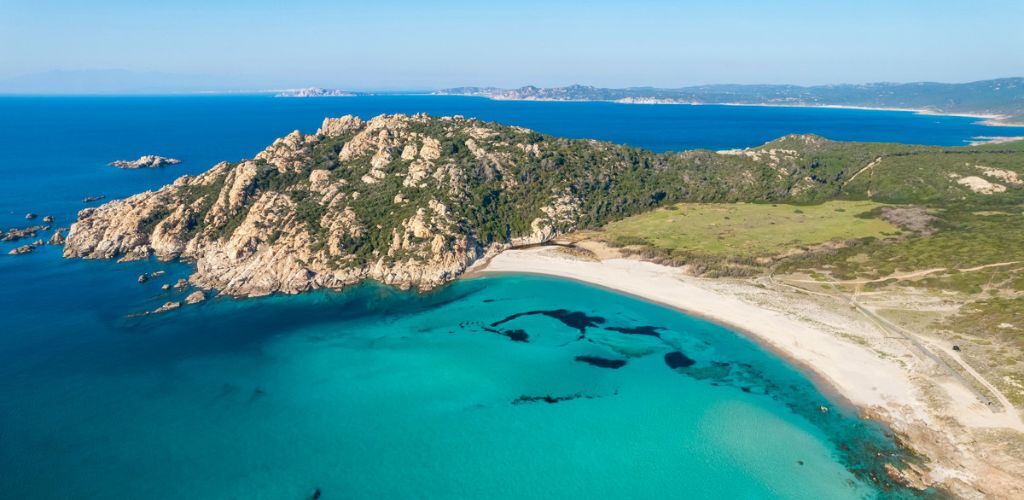Aerial view of turquoise bay and white sandy beach with rocky headland and green coastal scrub under clear blue sky