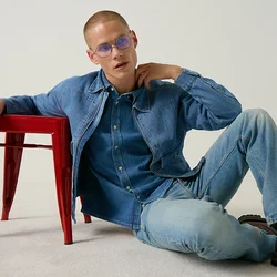 Uomo con giacca di jeans blu e jeans con scarpe marroni si appoggia a uno sgabello rosso contro uno sfondo chiaro e uniforme.