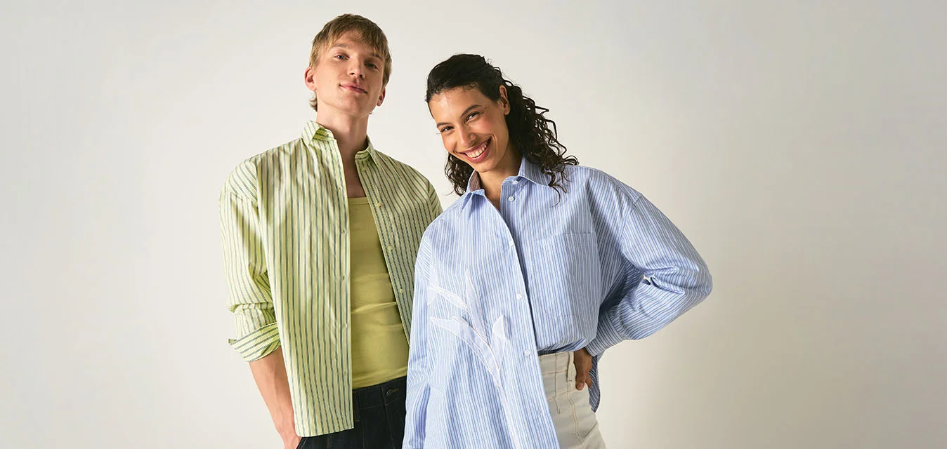 Due persone stanno sorridenti contro uno sfondo semplice, indossando camicie larghe a righe e pantaloni casual, posando con sicurezza.