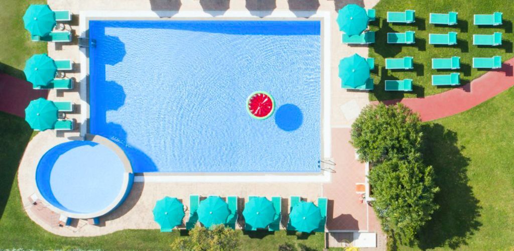 Aerial view of resort swimming pool with turquoise sun loungers and parasols, round kids' pool and a watermelon float in blue water