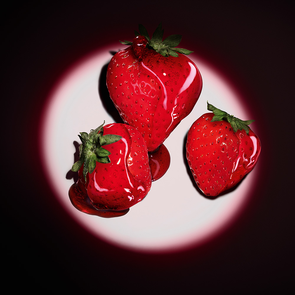 Tre fragole rosse fresche glassate con uno sciroppo rosso lucido sotto un riflettore circolare.