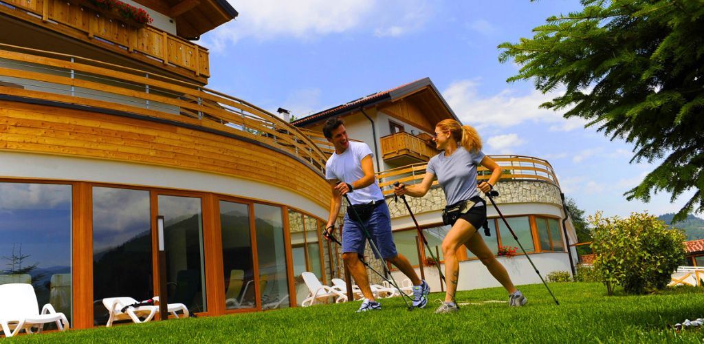 Two adults Nordic walking with poles on a grassy lawn in front of a wooden chalet-style hotel under a blue sky