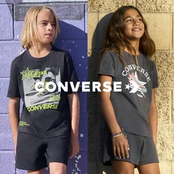 Due bambini che indossano magliette grafiche Converse e pantaloncini stanno contro pareti testurizzate, una viola e una beige, sorridenti e guardano altrove.