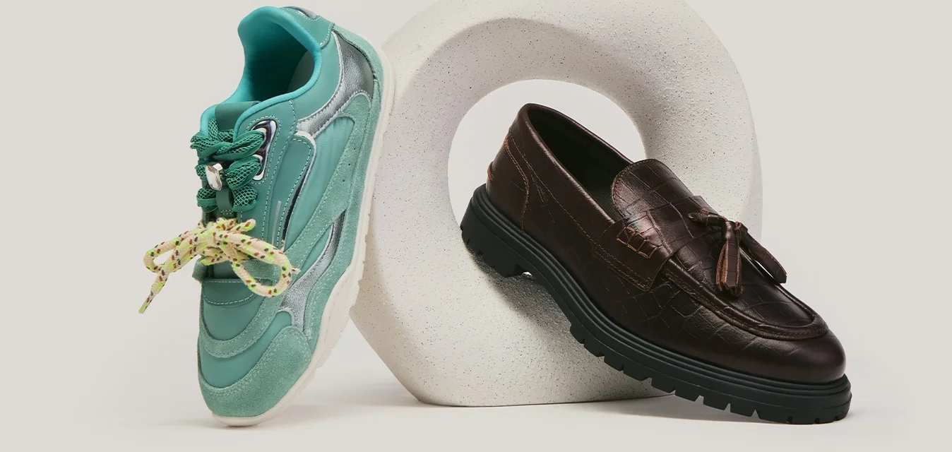 Sneaker verde con lacci gialli e mocassino in pelle marrone scuro esposti sopra e accanto a una scultura circolare bianca su sfondo grigio.