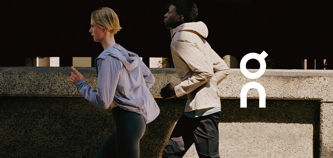 Due persone fanno jogging all'aperto indossando giacche sportive, leggings e scarpe da corsa su una strada urbana soleggiata.