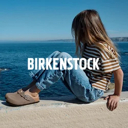 Ragazza con i capelli lunghi, che indossa una maglietta a righe e i jeans, seduta su un muro vicino al mare, guarda verso l'acqua sotto un cielo limpido.