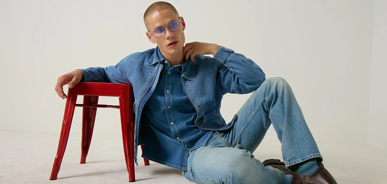 Uomo con giacca di jeans blu e jeans con scarpe marroni si appoggia a uno sgabello rosso contro uno sfondo chiaro e uniforme.