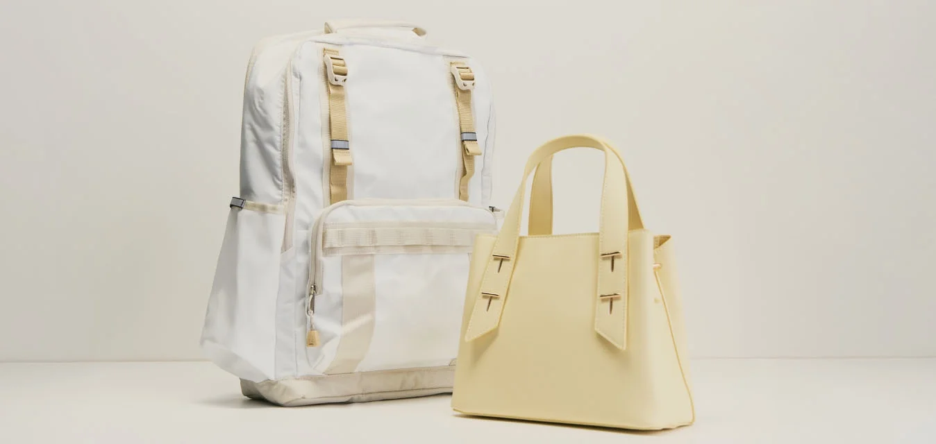 Zaino bianco con spallacci beige accanto a una piccola borsa beige su uno sfondo chiaro e uniforme, entrambi in piedi.