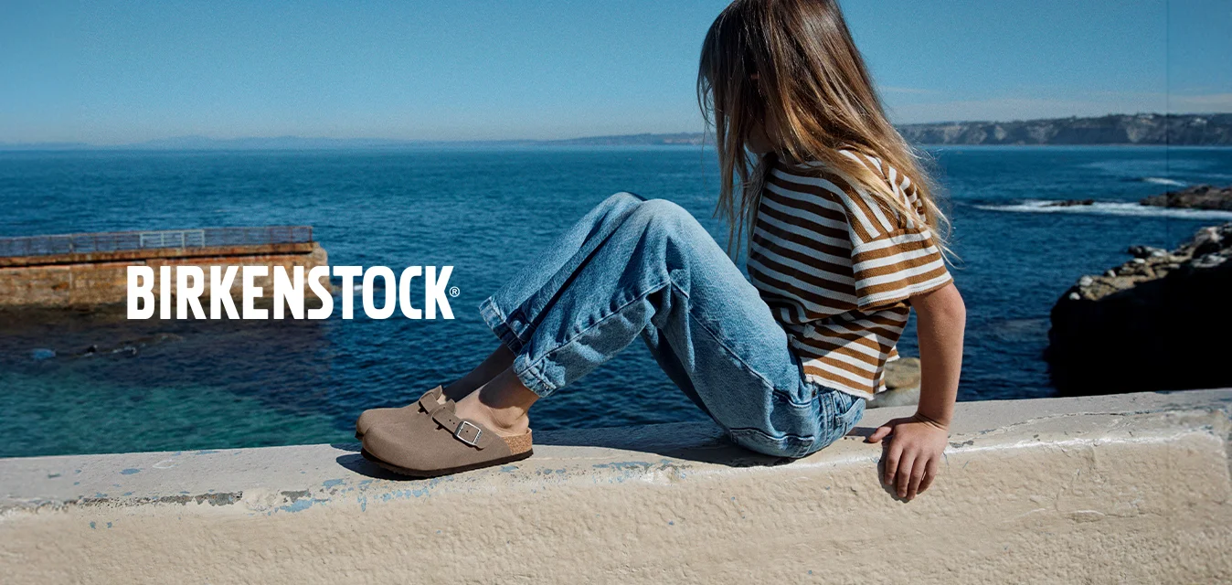 Ragazza con i capelli lunghi, che indossa una maglietta a righe e i jeans, seduta su un muro vicino al mare, guarda verso l'acqua sotto un cielo limpido.