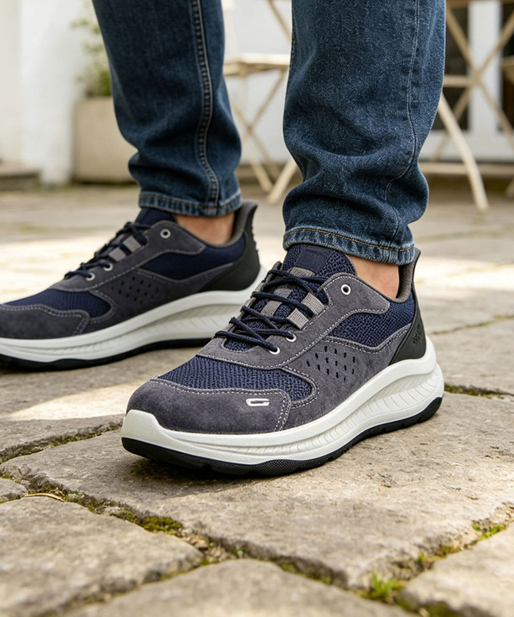 Pair of navy and grey men's walking trainers with cushioned white sole on cobblestone path, wearer in blue jeans