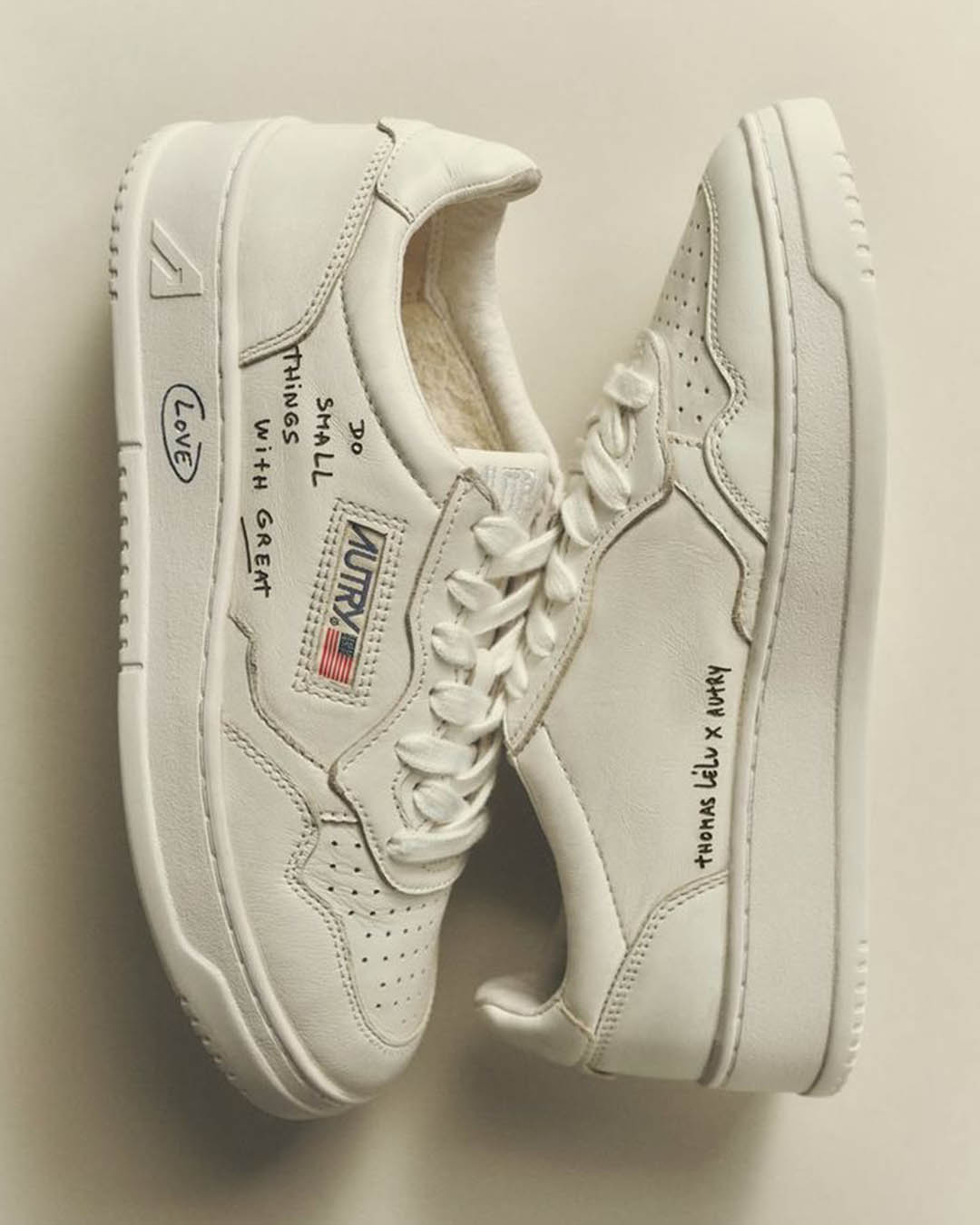 Pair of white Autry leather trainers with handwritten black text and small US flag logo, side-by-side on pale background