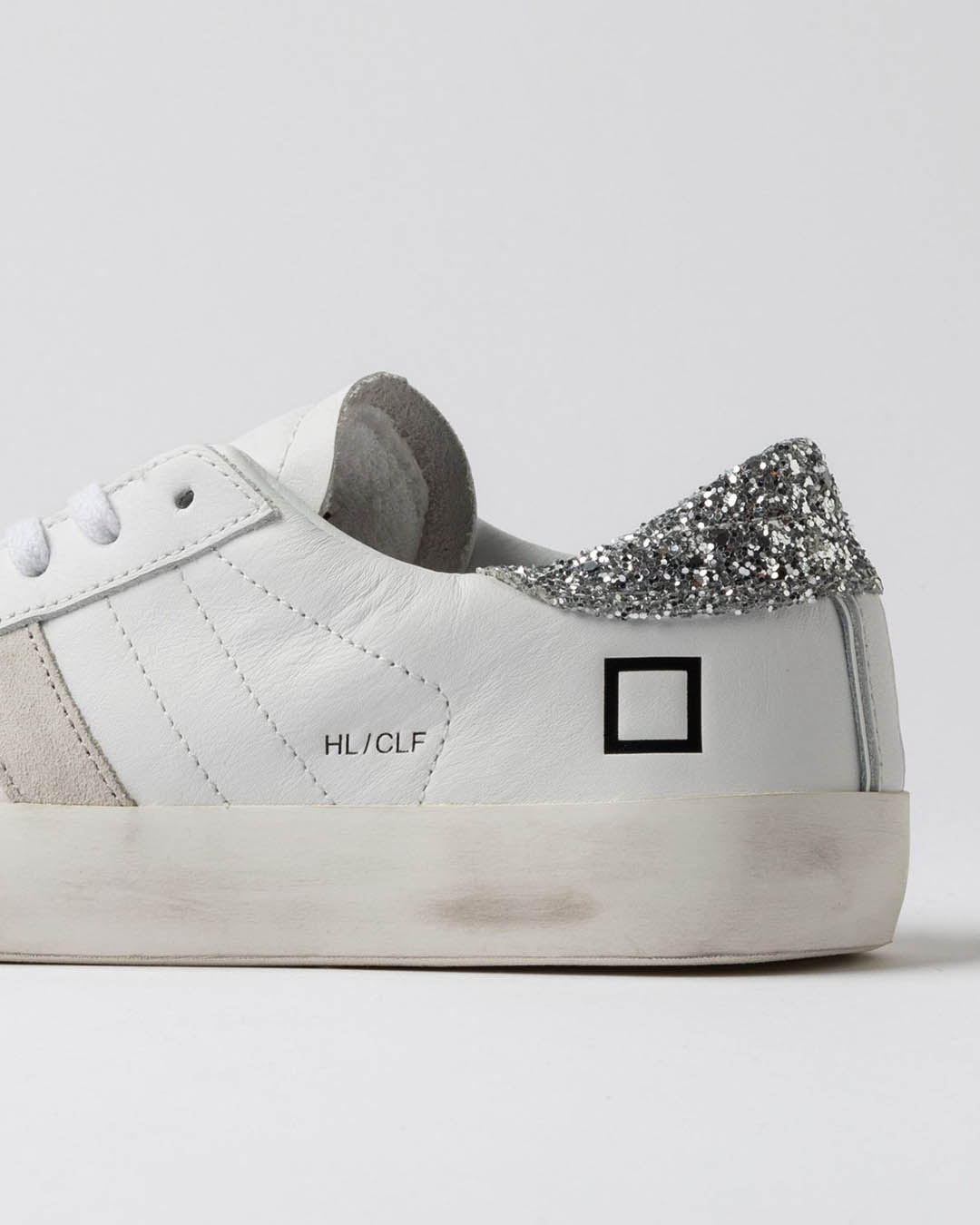 White leather trainer close-up showing silver glitter heel patch, black square logo and 'HL/CLF' text on slightly scuffed sole