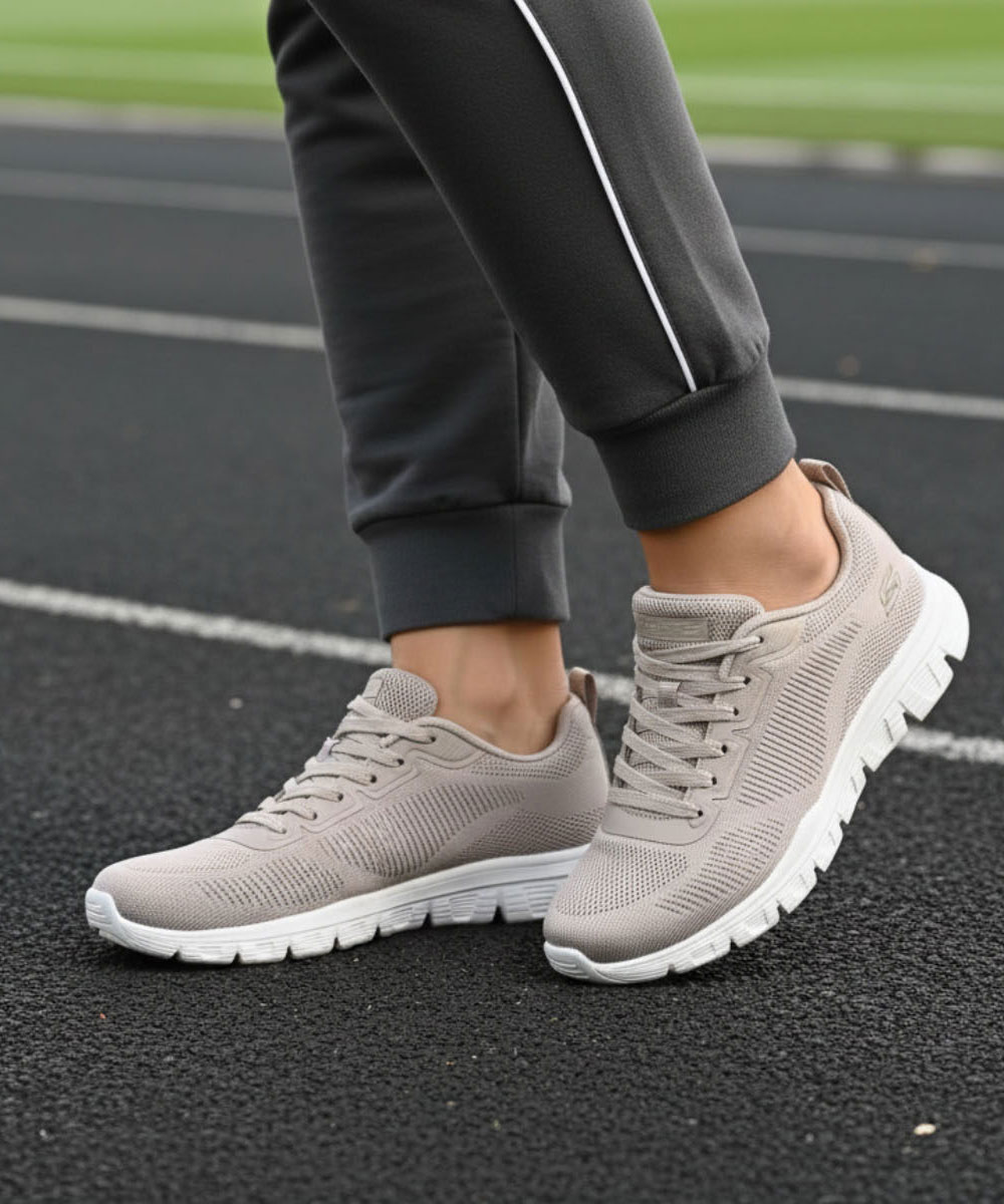 Pair of beige knitted trainers with white cushioned soles on an athletics track, worn with dark jogging bottoms