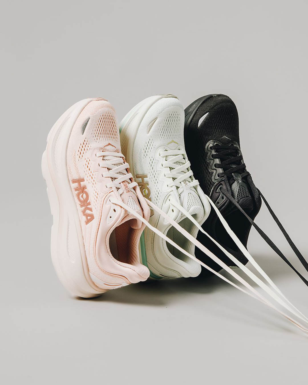 Three Hoka running trainers (pink, white, black) with extended laces arranged side-by-side on a pale grey background