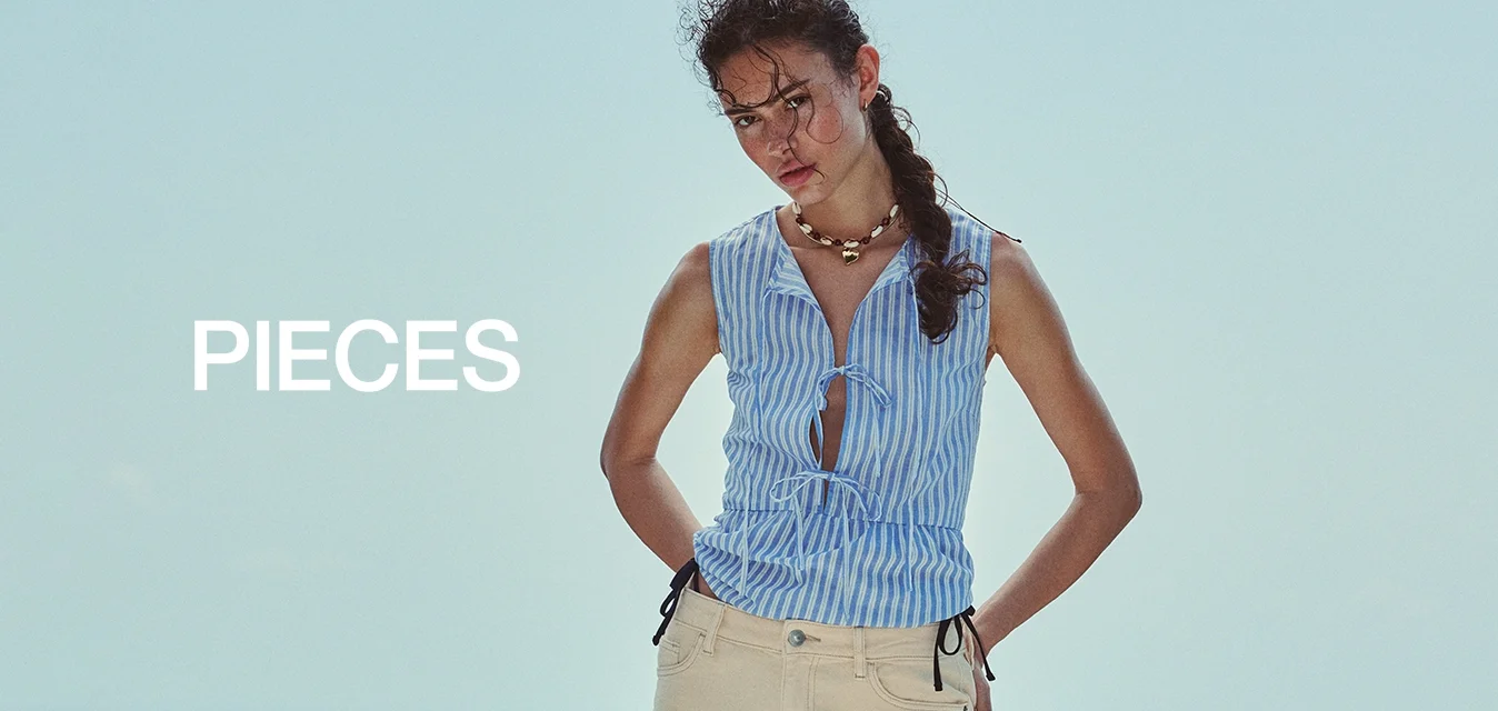 Donna con capelli intrecciati in piedi su una piattaforma in legno vicino al mare, indossa un top blu a righe senza maniche, pantaloncini beige al ginocchio e infradito nere.