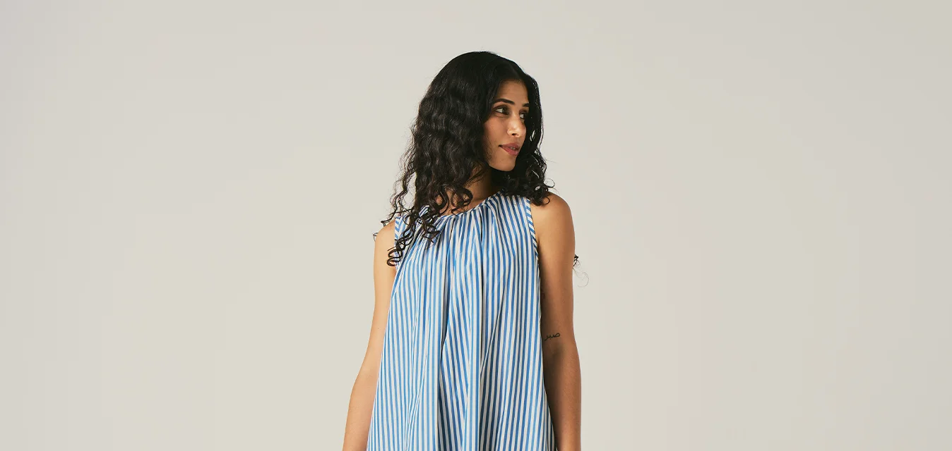 Donna con lunghi capelli ricci che indossa un vestito lungo senza maniche a righe verticali blu e bianche, in piedi davanti a uno sfondo chiaro uniforme.