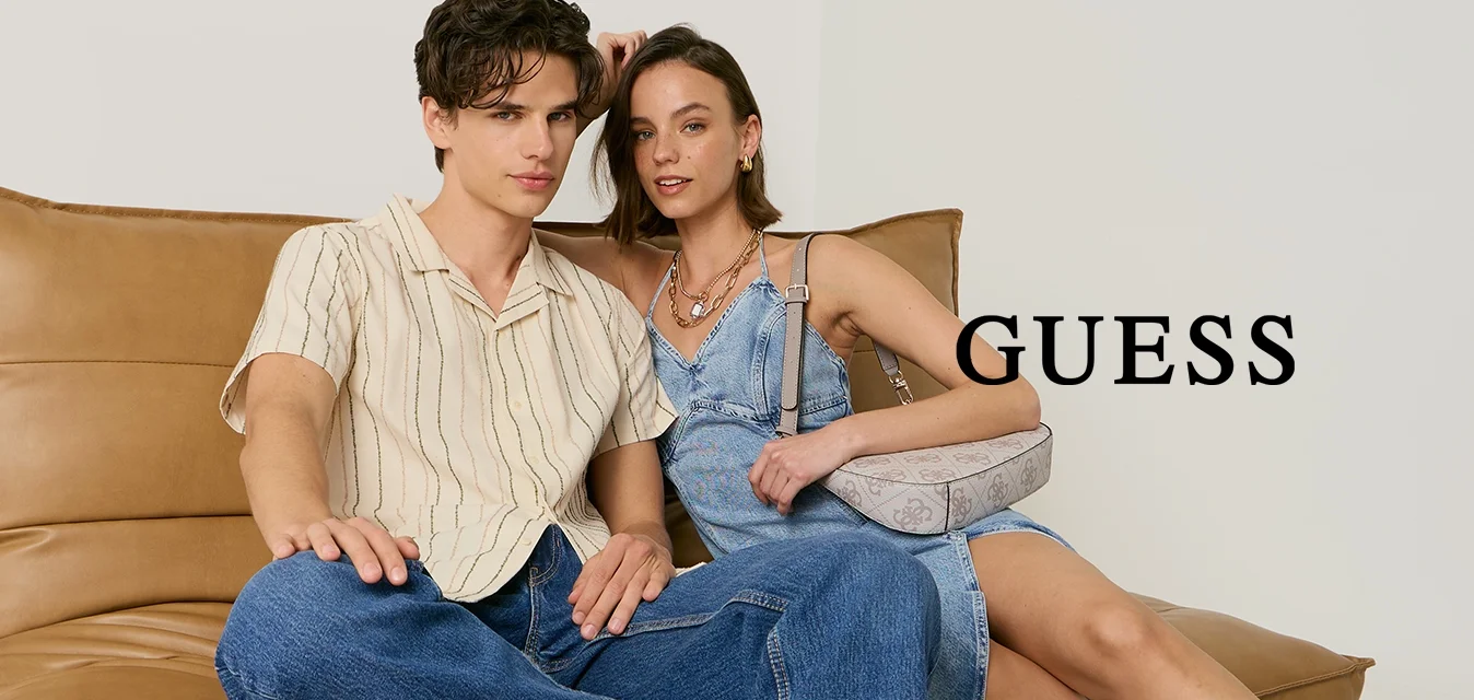 Giovane uomo con camicia a righe e jeans e giovane donna con vestito blu e tacchi neri seduti su un divano in pelle color cuoio contro uno sfondo semplice.