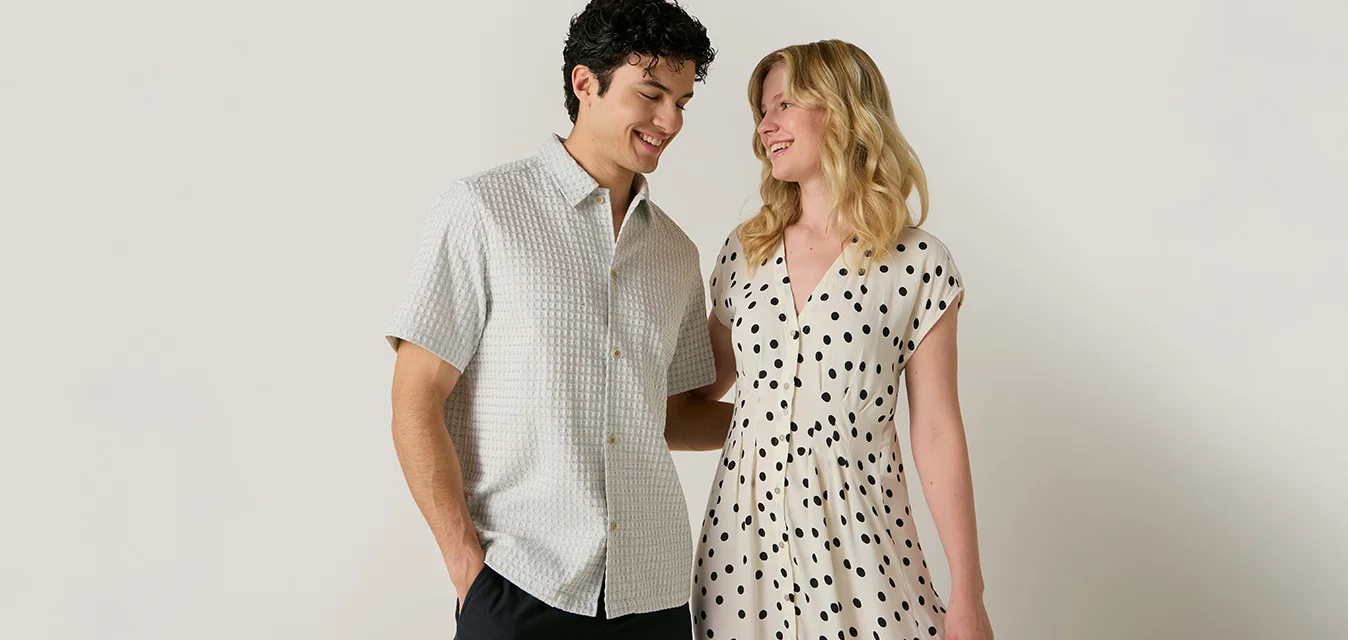 Un uomo in camicia a maniche corte e pantaloncini neri sta accanto a una donna con un vestito bianco con puntini neri, entrambi sorridono e si guardano.