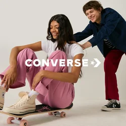 Giovane donna con pantaloni rosa seduta su uno skateboard mentre un uomo sorridente la spinge da dietro in uno spazio studio bianco.