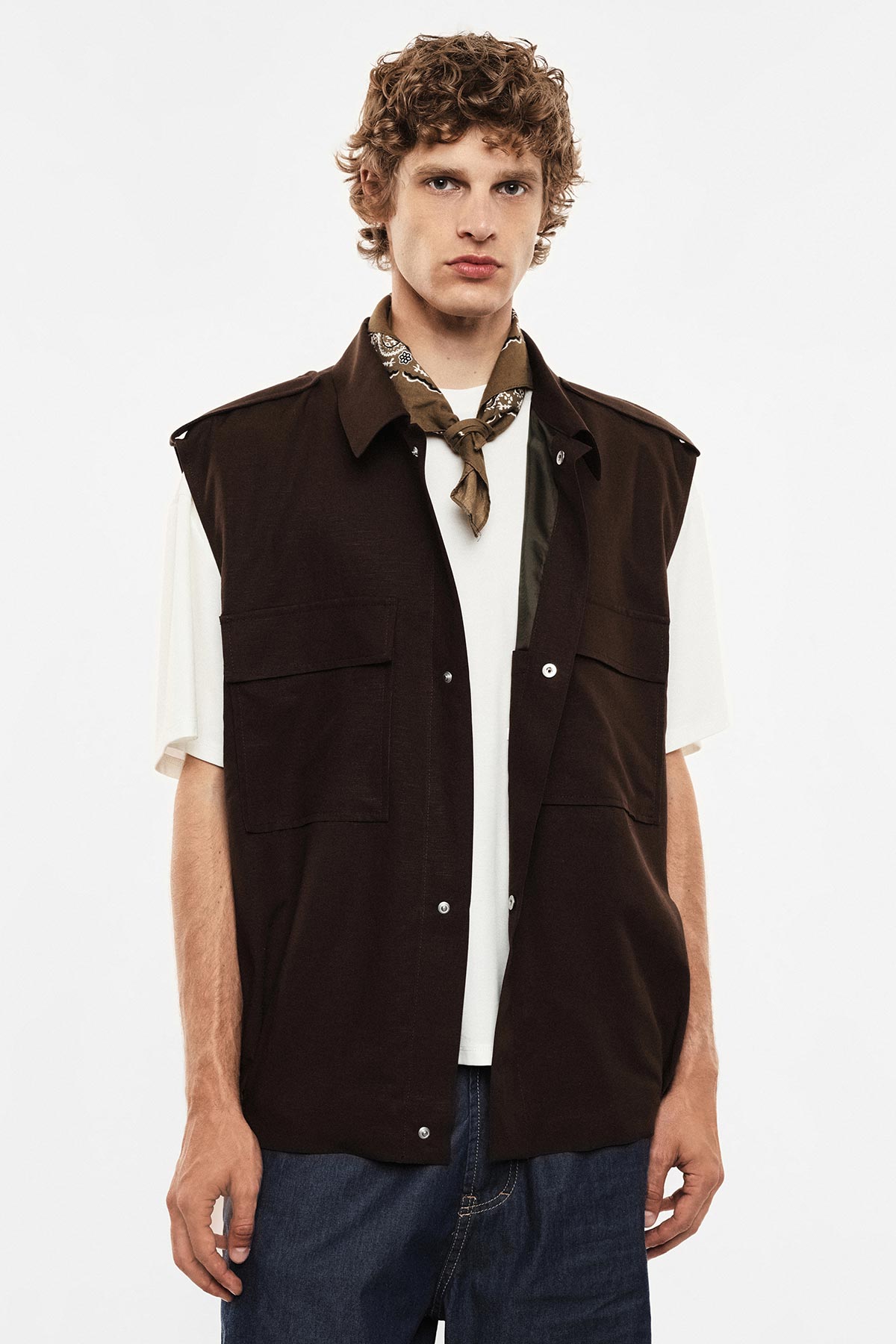 Man in dark brown sleeveless jacket over white T-shirt, bandana neckerchief and blue jeans on plain studio background