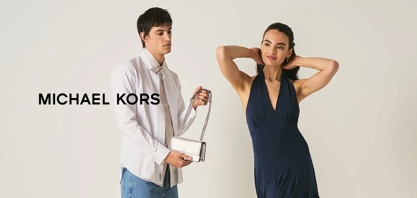 Giovane donna in abito blu navy posa mentre un uomo con camicia bianca e jeans tiene una piccola borsa bianca in una stanza semplice.